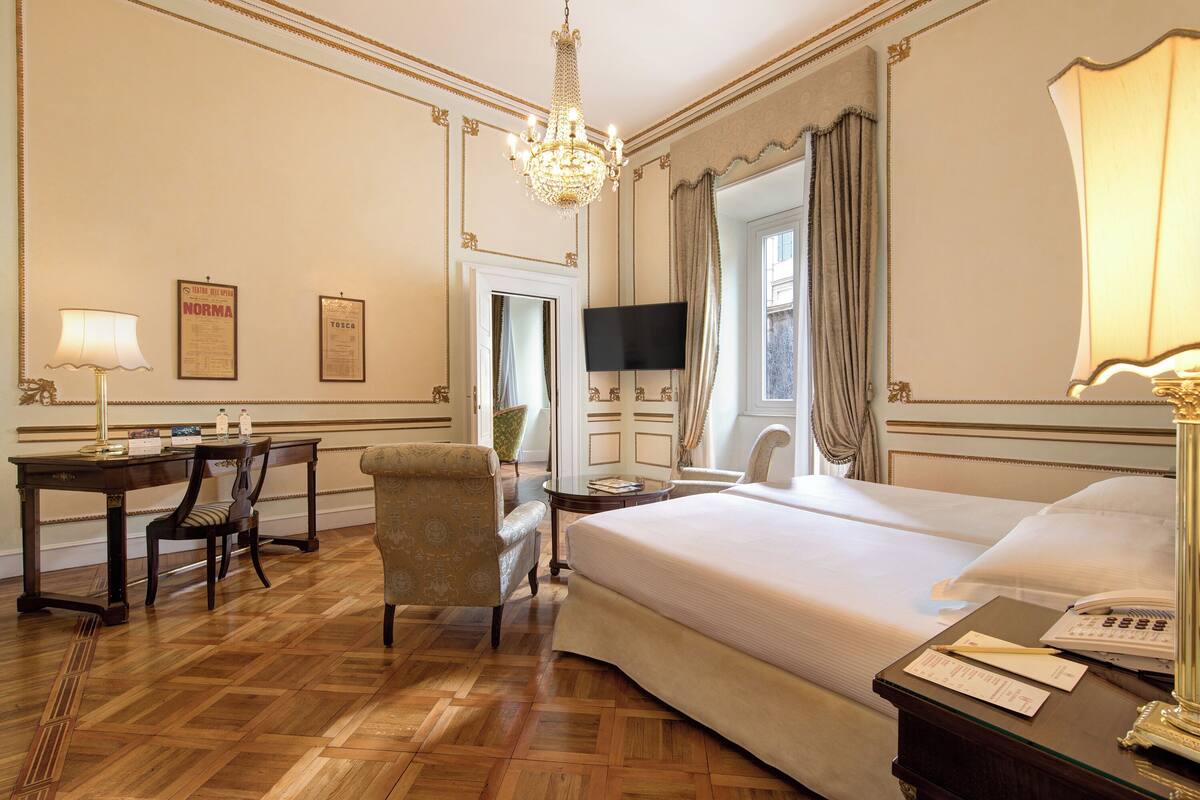 A hotel room with a large bed, a desk with a lamp, a chair, a television, and a chandelier.