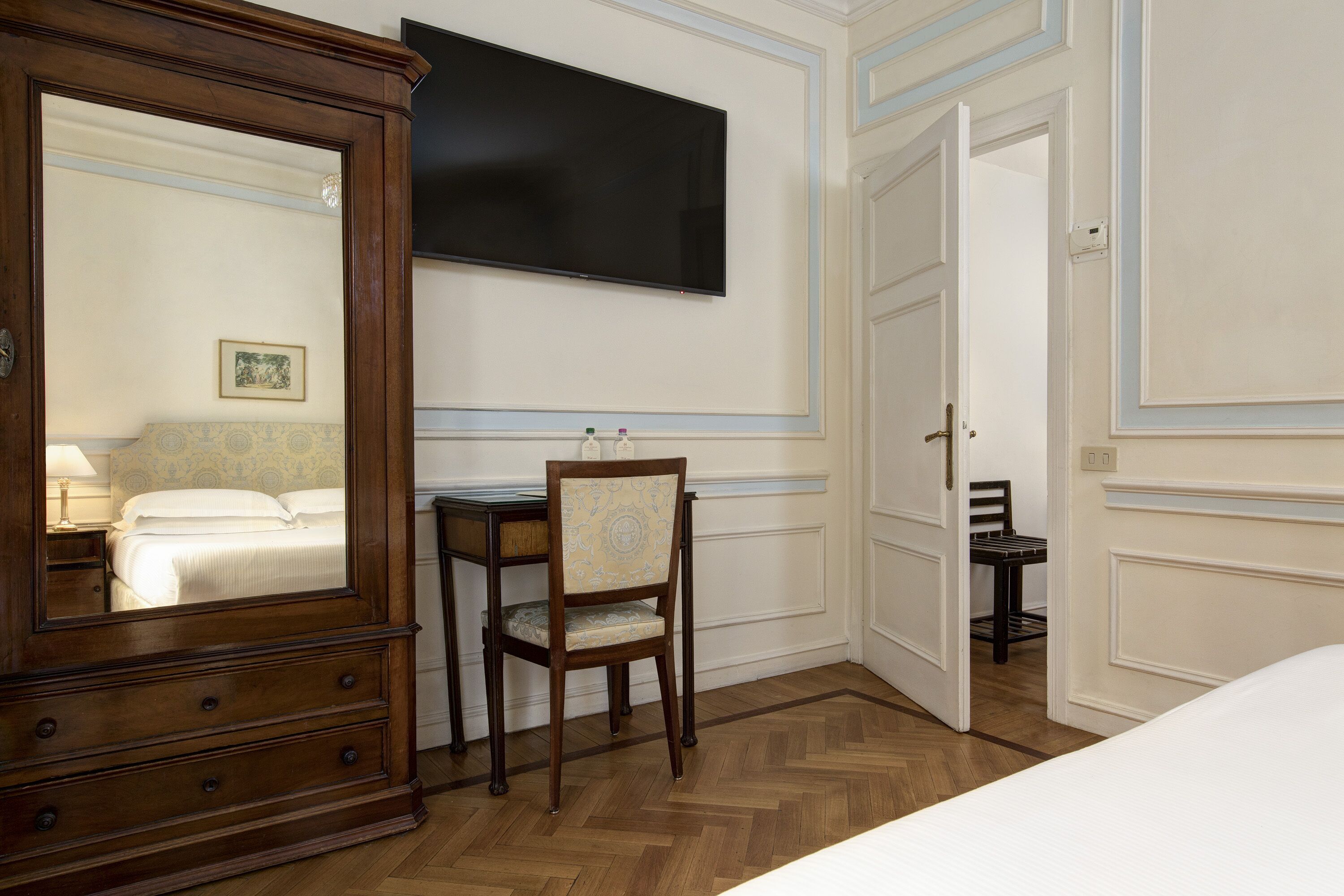 A hotel room with a wooden dresser, a desk with a chair, a flat-screen TV mounted on the wall, and a bed with white bedding.