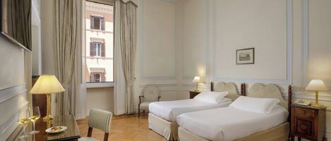 A hotel room with two beds, a desk with a lamp, a chair, and a view of a building through the window.