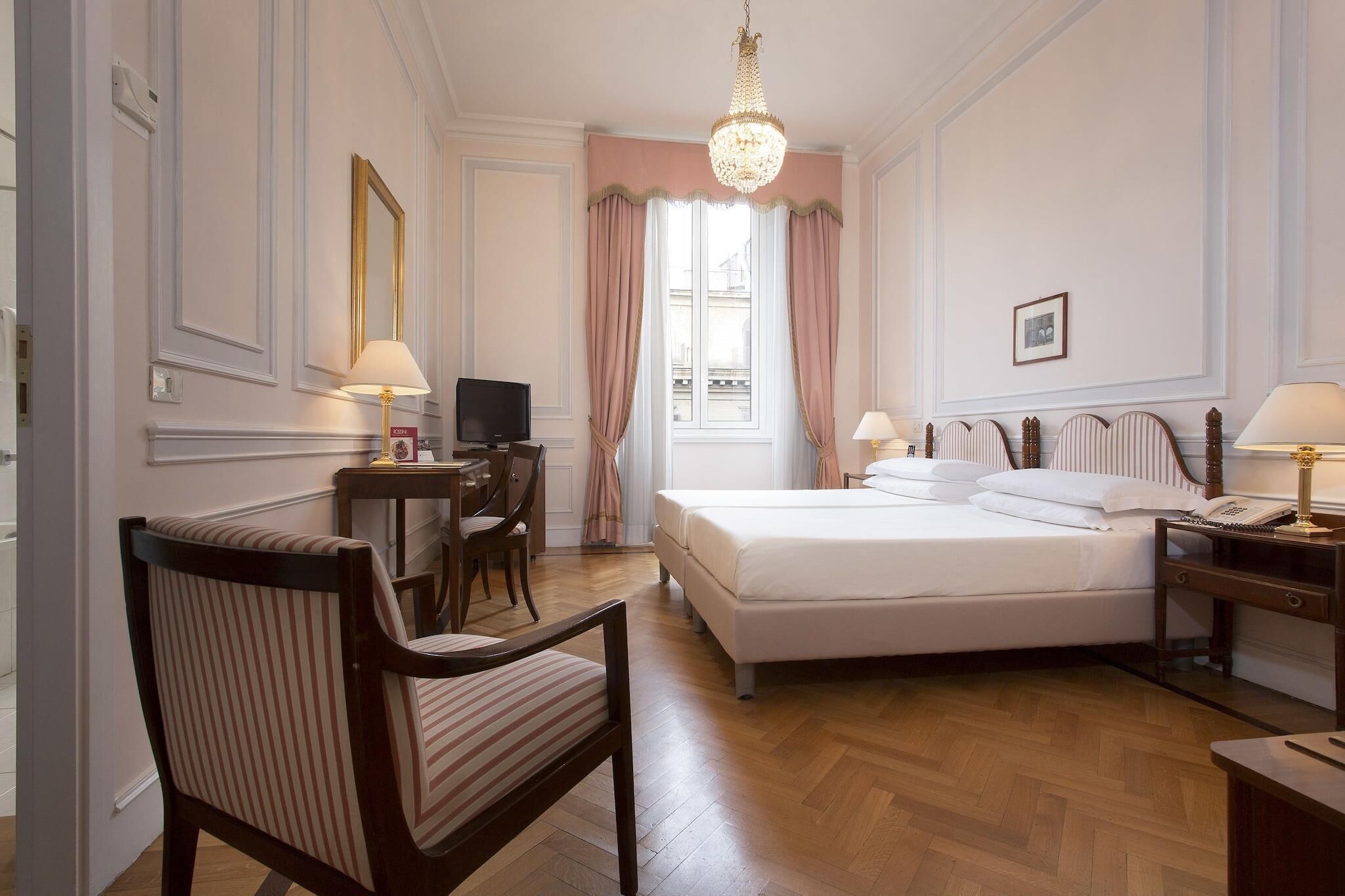 Habitación de hotel con cama, escritorio con computadora, silla, lámpara de escritorio, mesita de noche con lámpara y candelabro.