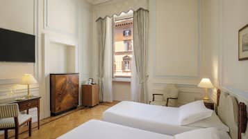 Una camera d'albergo con due letti, una scrivania con lampada, una sedia, un armadio in legno e vista su un edificio attraverso una finestra con tende.