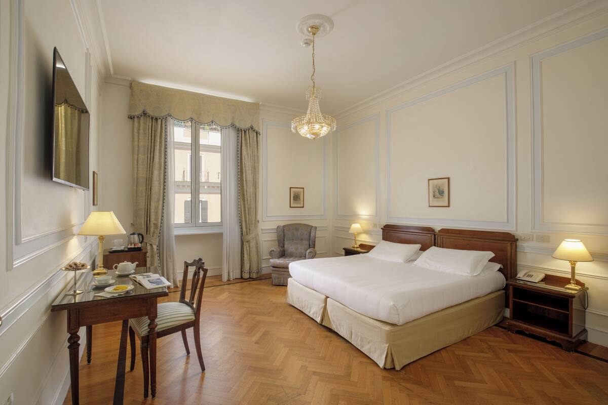 A hotel room with a large bed, a desk with a chair, a chair, a nightstand with a lamp, and a chandelier.