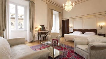 A hotel room with a large bed, two armchairs, a desk with a lamp, and a chandelier.