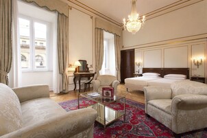 A hotel room with a large bed, two armchairs, a desk with a lamp, and a chandelier.