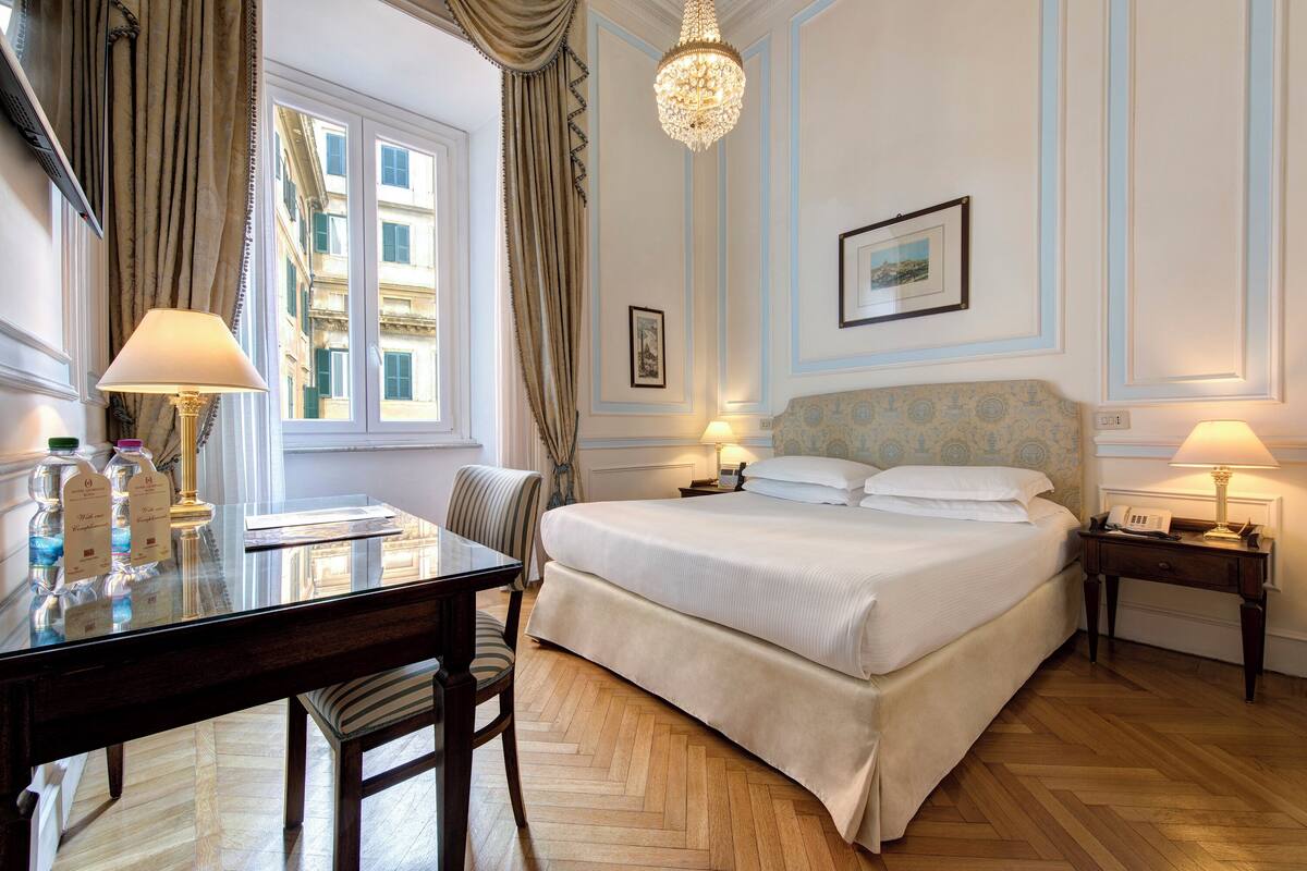 A hotel room with a large bed, two bedside tables with lamps, a desk with a chair, a chandelier, and a window with curtains.