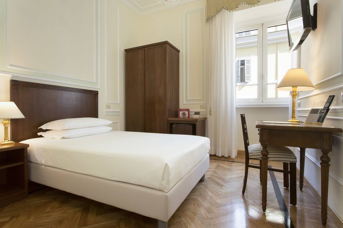 A hotel room with a bed, a desk, a chair, a wardrobe, and a window with curtains.