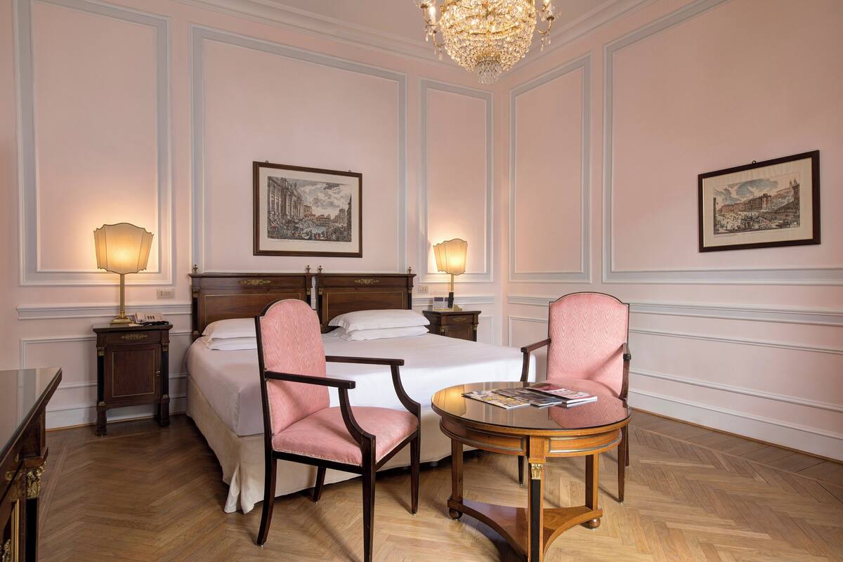 A hotel room with a large bed, two bedside lamps, a wooden chair, a small round table, and two framed pictures on the walls.