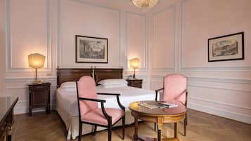 Hotelkamer met een groot bed, twee bedlampen, een houten stoel, een klein rond tafeltje en twee foto's in lijsten aan de muur.