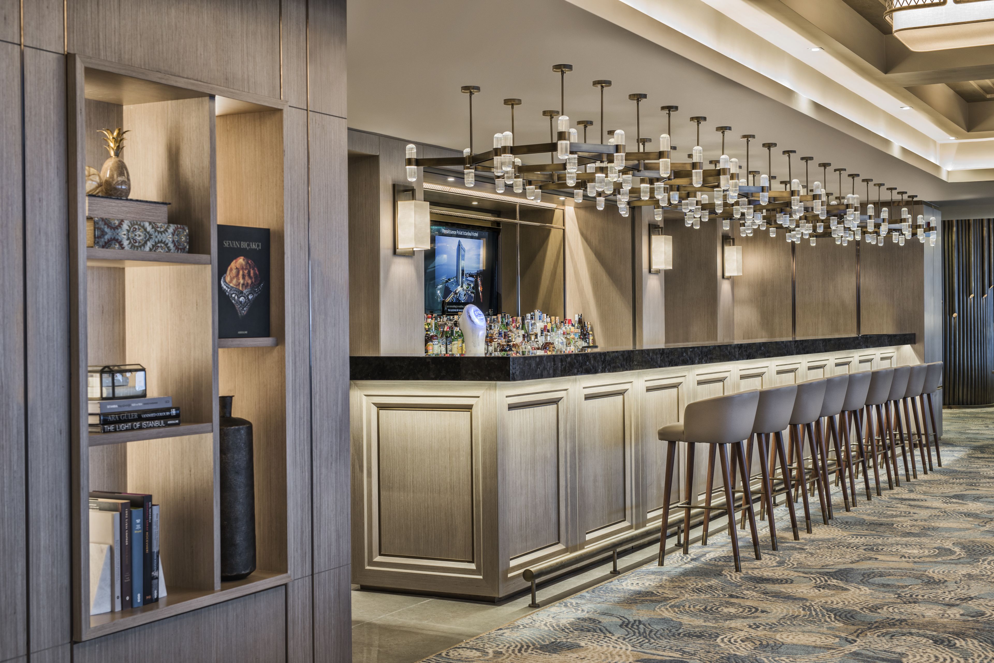 A bar area with a long counter, multiple stools, and a modern chandelier.