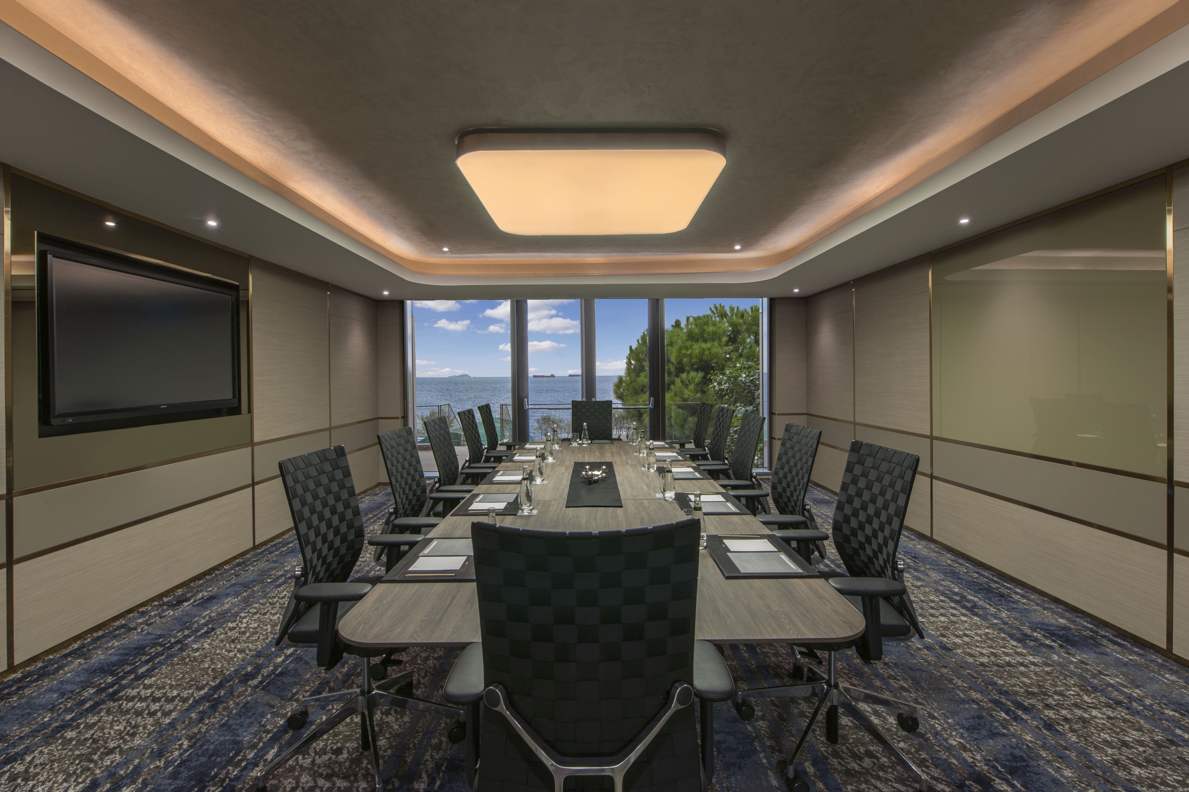 Un salón de conferencias con una mesa larga, sillas, una pantalla de proyector y grandes ventanales con vista al mar.