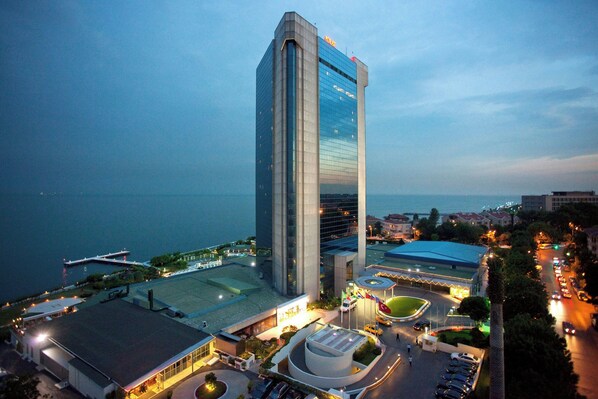 A modern high-rise hotel with a prominent vertical design, a parking area, and a waterfront view.