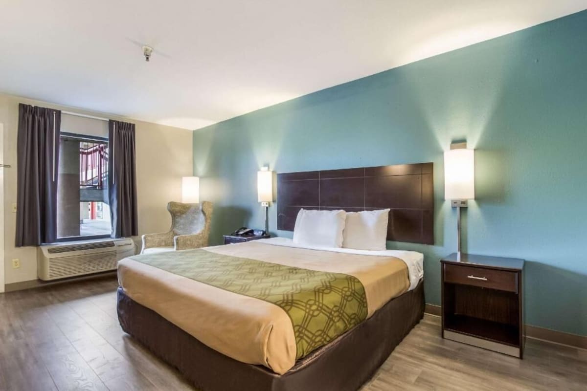 A hotel room with a large bed, bedside lamps, a nightstand, a chair, and a window with curtains.