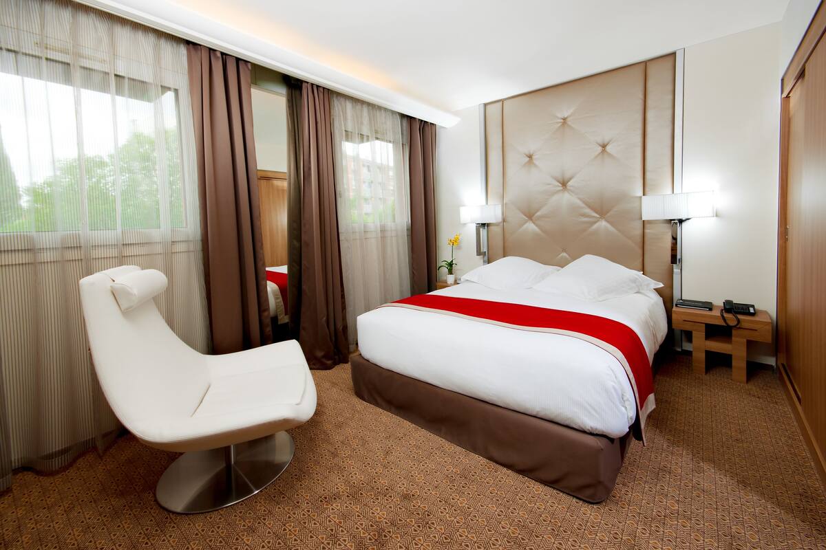 A hotel room with a large bed, a white armchair, a bedside table with a lamp, and a view of the outdoors through sheer curtains.