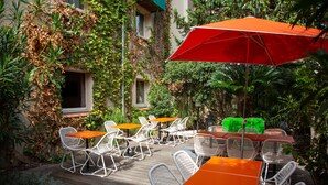 An outdoor patio with white wicker chairs and orange tables, surrounded by lush greenery and a red umbrella. - Hôtel Mermoz (Toulouse)