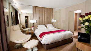 A hotel room with a large bed, a sitting area with a chair and ottoman, a desk with a lamp, and a mirror. - Hôtel Mermoz (Toulouse)