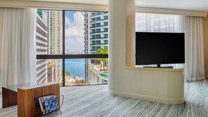 A hotel room with a large window, a flat-screen TV, a wooden desk, and a magazine rack.