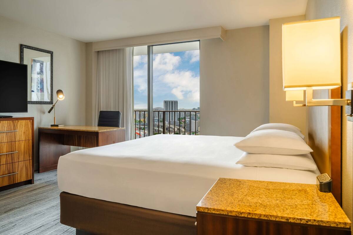 A hotel room with a large bed, a desk, a television, and a view of the cityscape through the window.