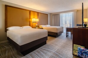 A hotel room with two beds, a desk, a lamp, and a view of the outdoors.