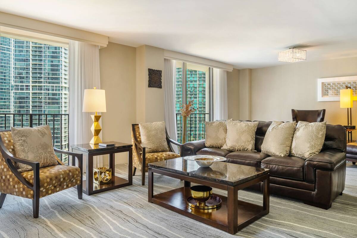 A modern living room with a leather sofa, two armchairs, a coffee table, and a view of a high-rise building through large windows.