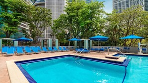 A swimming pool with blue lounge chairs and umbrellas, surrounded by trees and modern buildings.