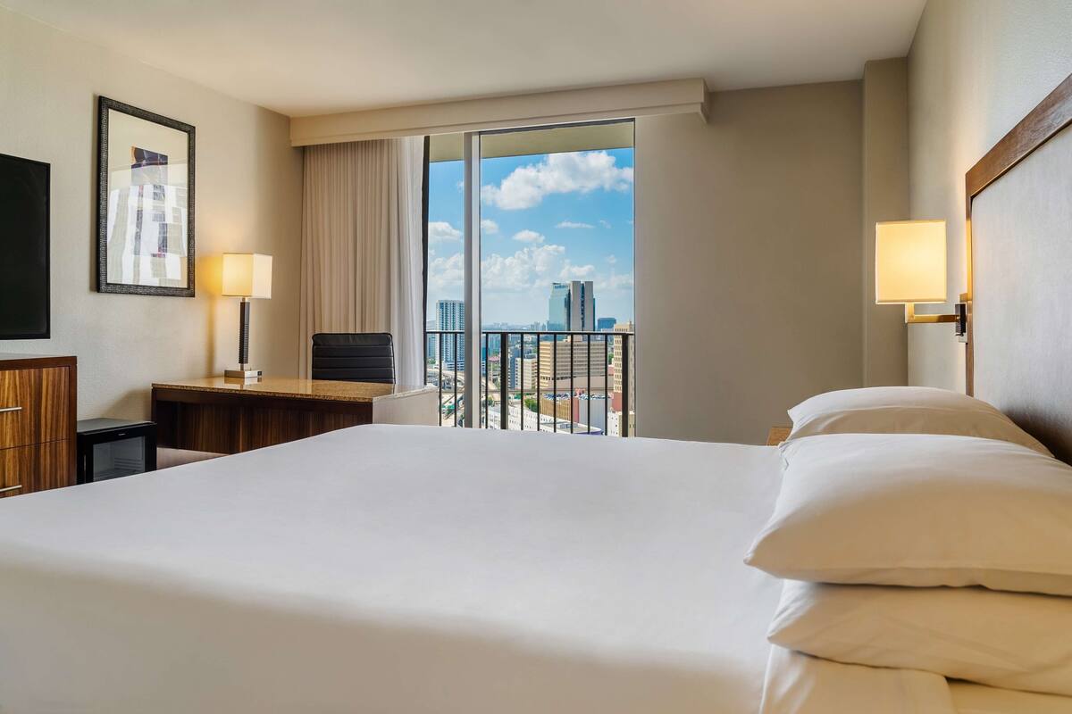 A hotel room with a large bed, a desk with a chair, a TV, and a view of the cityscape through a large window.