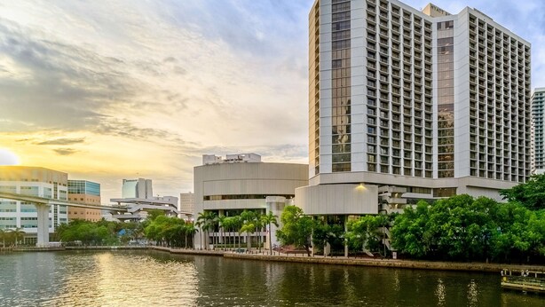 A modern hotel with a prominent corner building, a waterfront view, and a bridge in the background.