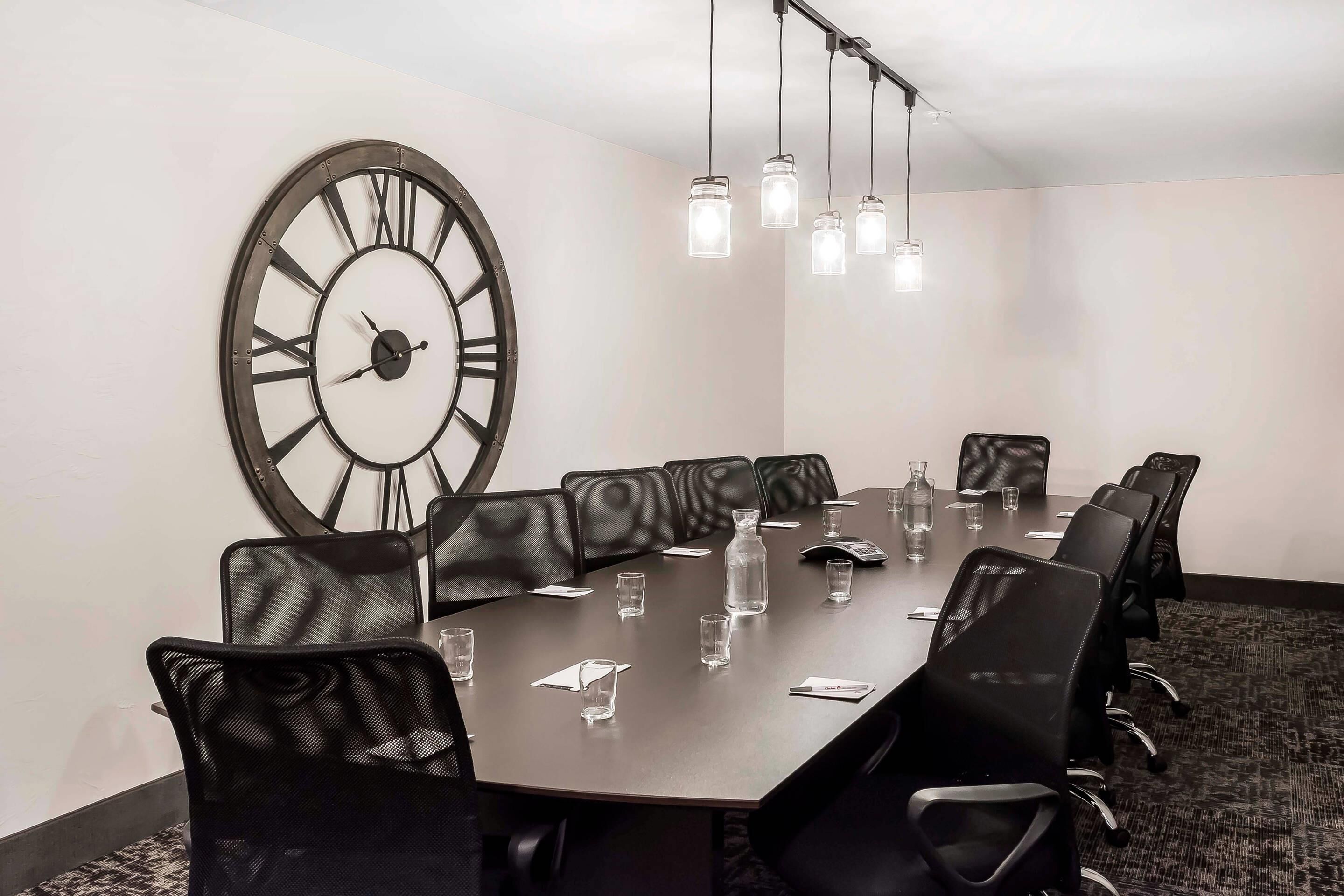 A conference room with a large clock, black leather chairs, a long table, and hanging lights.
