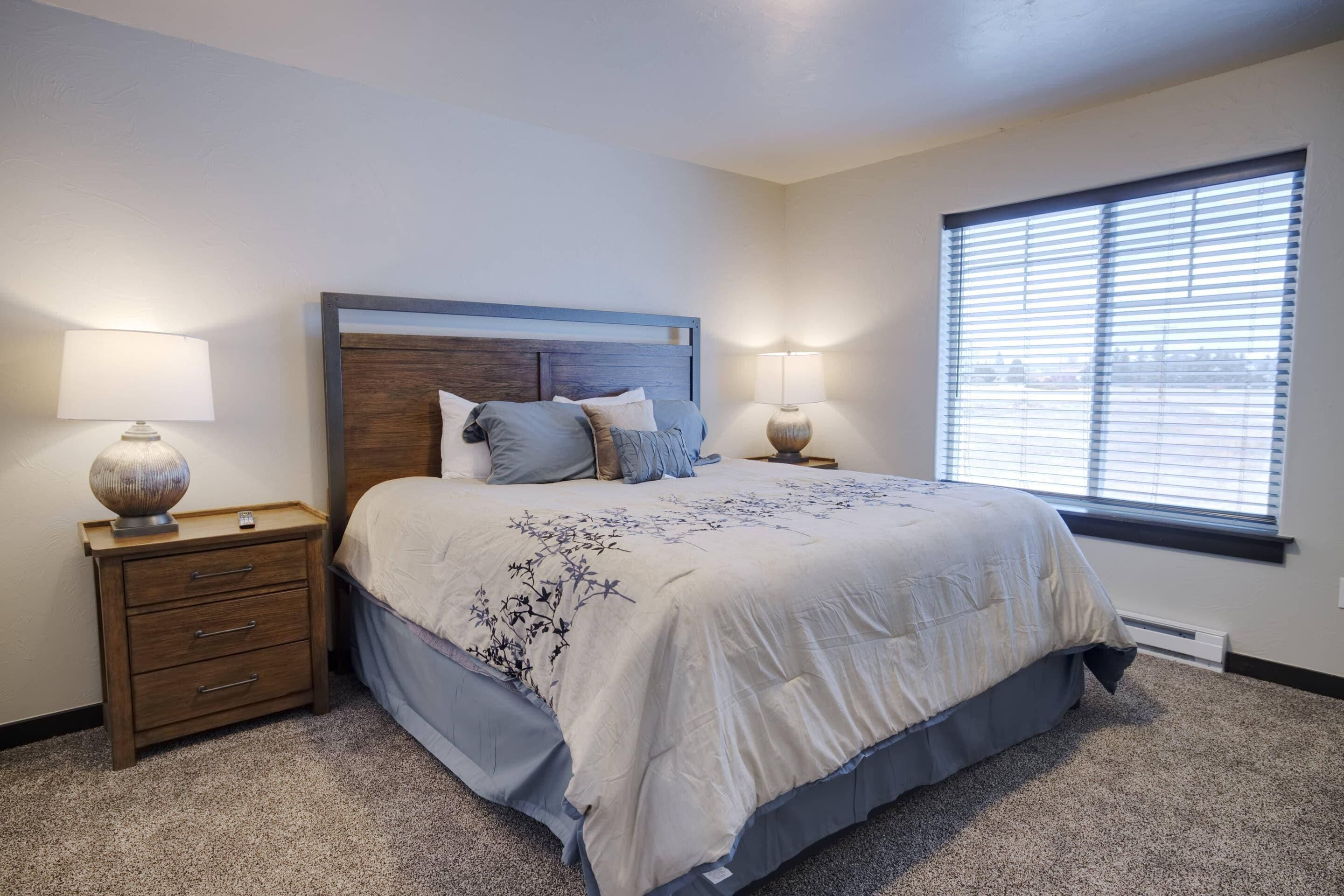 A bedroom with a large bed, two bedside tables with lamps, a window with blinds, and a carpeted floor.