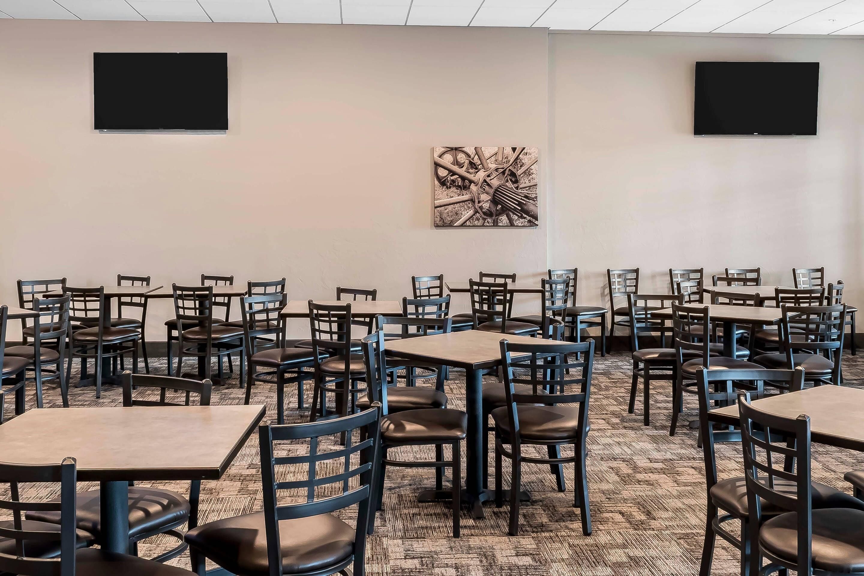 A conference room with multiple tables and chairs, two large screens on the walls, and a piece of abstract art.