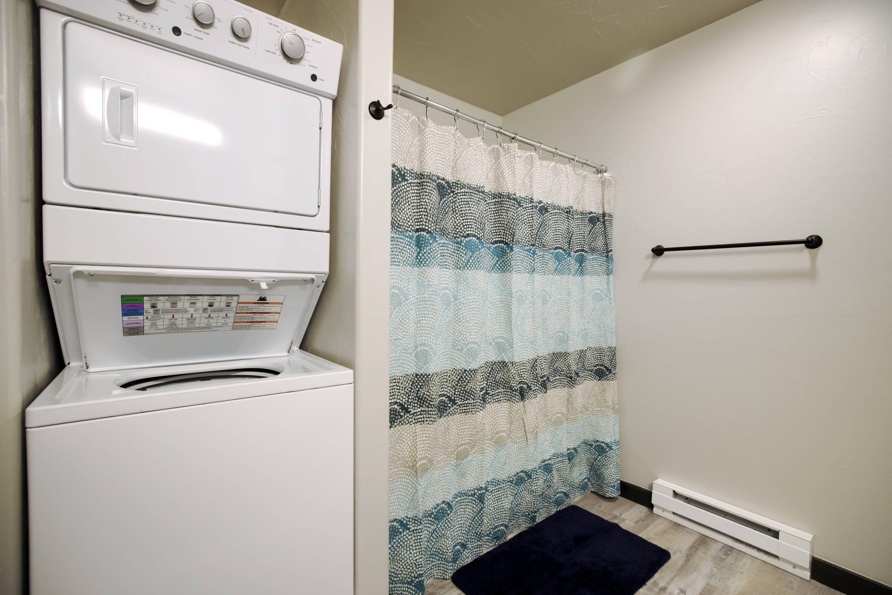 Een wasserij met een gestapelde wasmachine en droger, een decoratieve douchegordijn, een zwarte handdoekhouder en een witte radiator.