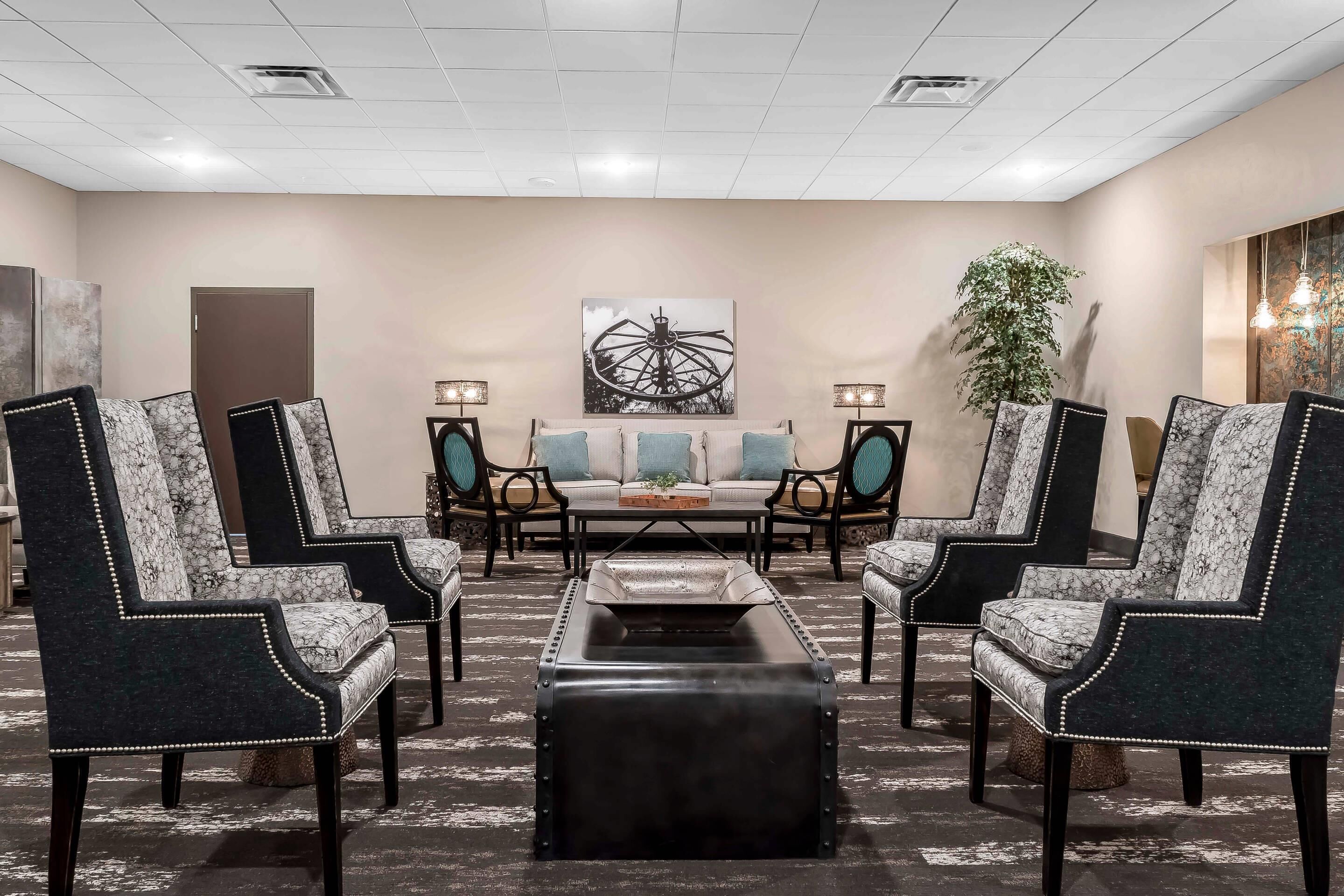 A hotel lobby with a central seating area featuring a sofa set, two armchairs, and a coffee table. There is a large abstract painting on the wall, a potted plant, and a door in the background.