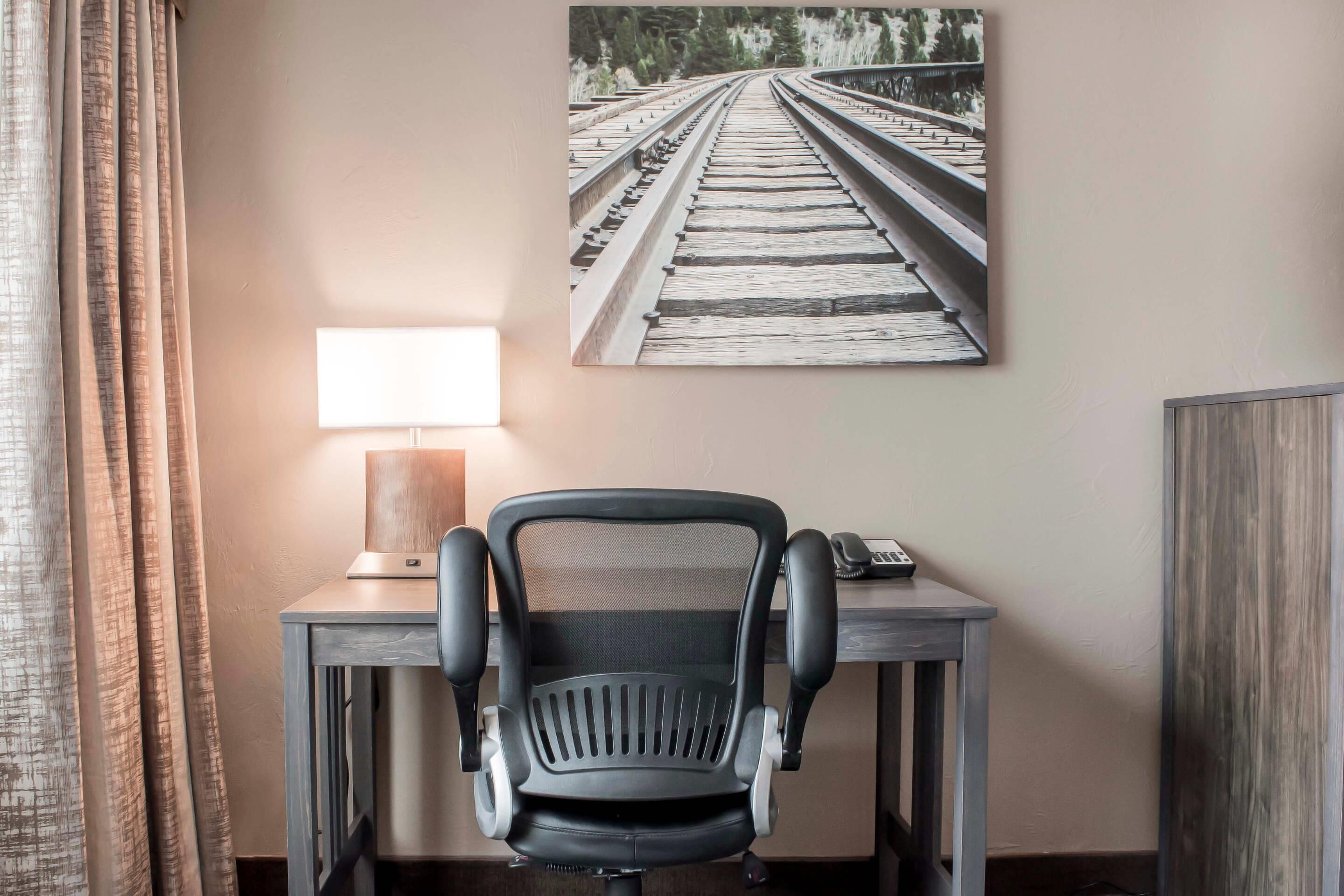A hotel room with a desk, a chair, a lamp, and a painting of train tracks on the wall.