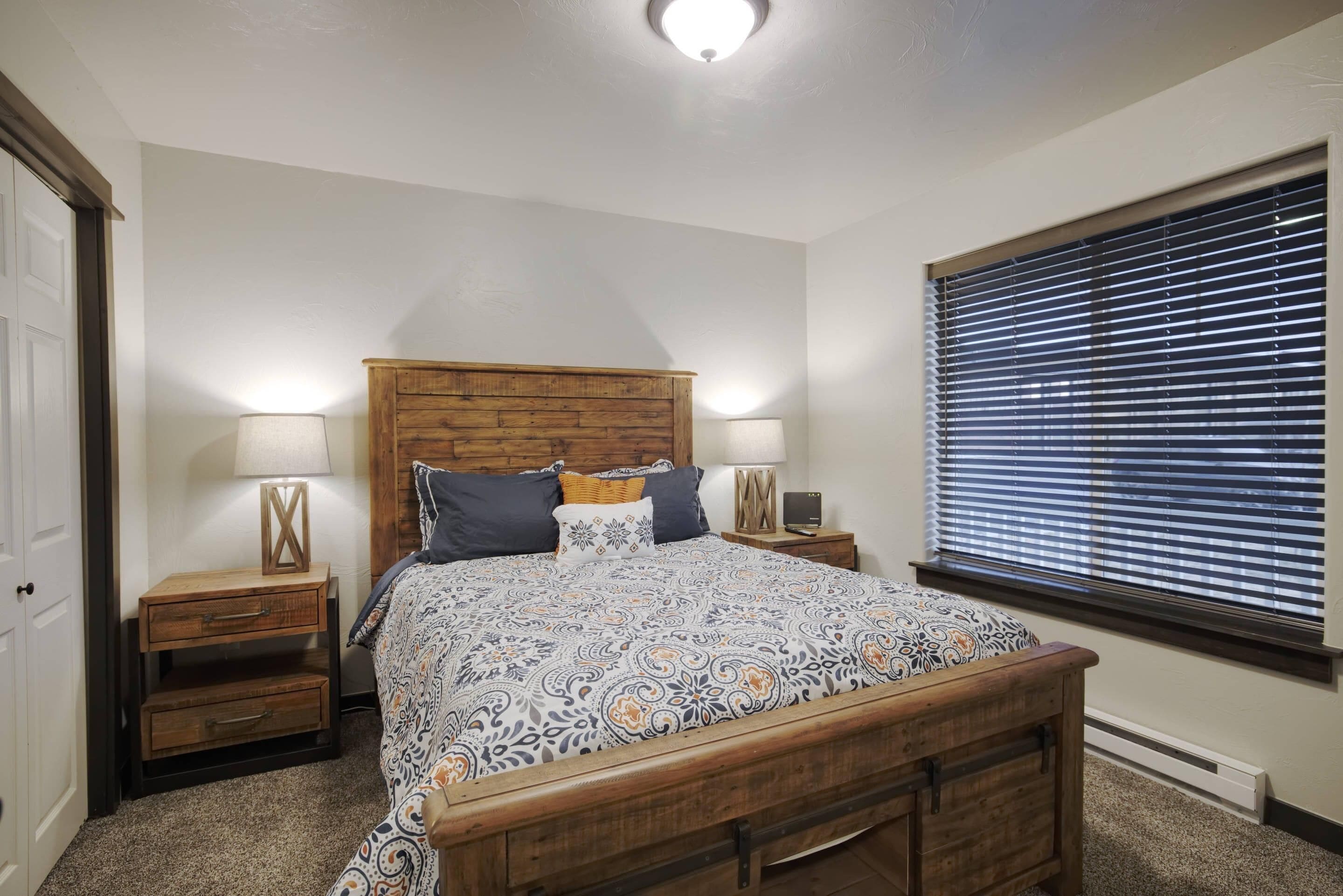 A bedroom with a wooden headboard, a patterned bedspread, two bedside tables with lamps, and a window with blinds.