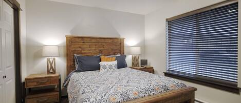 A bedroom with a wooden headboard, a patterned bedspread, two bedside tables with lamps, and a window with blinds.