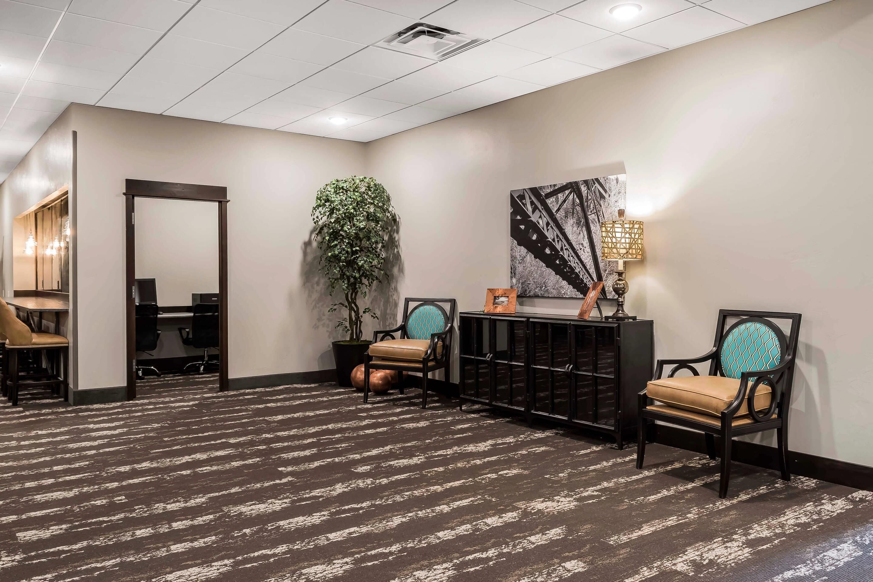 A hotel lobby with a patterned carpet, two upholstered chairs, a desk, a large potted plant, and a wall-mounted lamp.