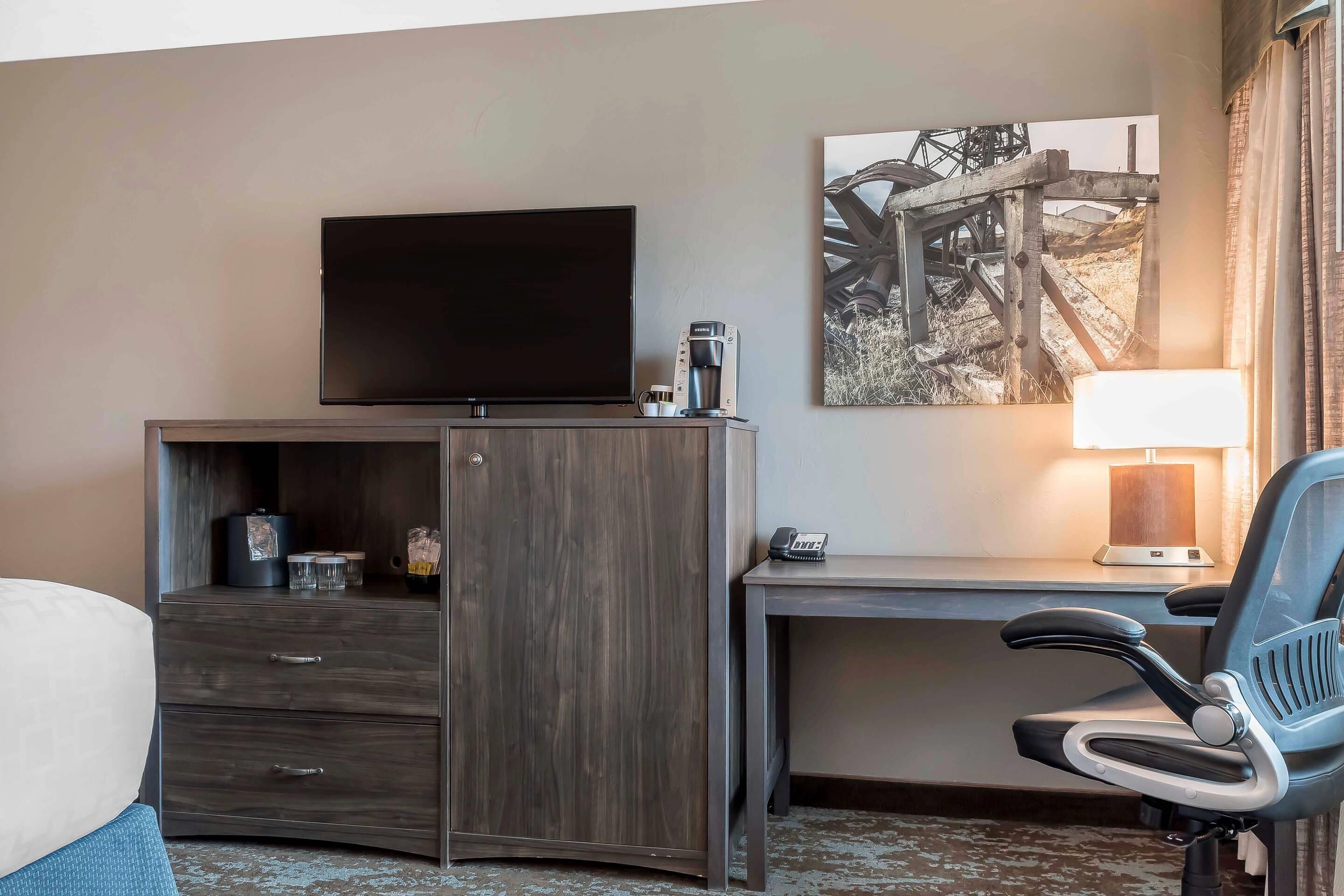 A hotel room with a TV on a wooden cabinet, a desk with a lamp, a chair, and a bed.