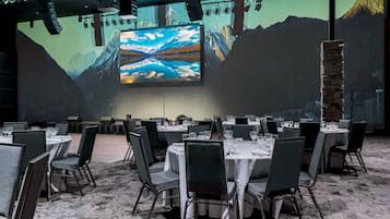 Uma sala de conferência com mesas redondas, cadeiras e uma tela grande exibindo uma paisagem montanhosa.