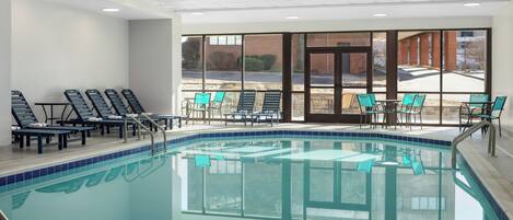 Una piscina interior con paredes de vidrio transparente, varias sillas de descanso y mesas.