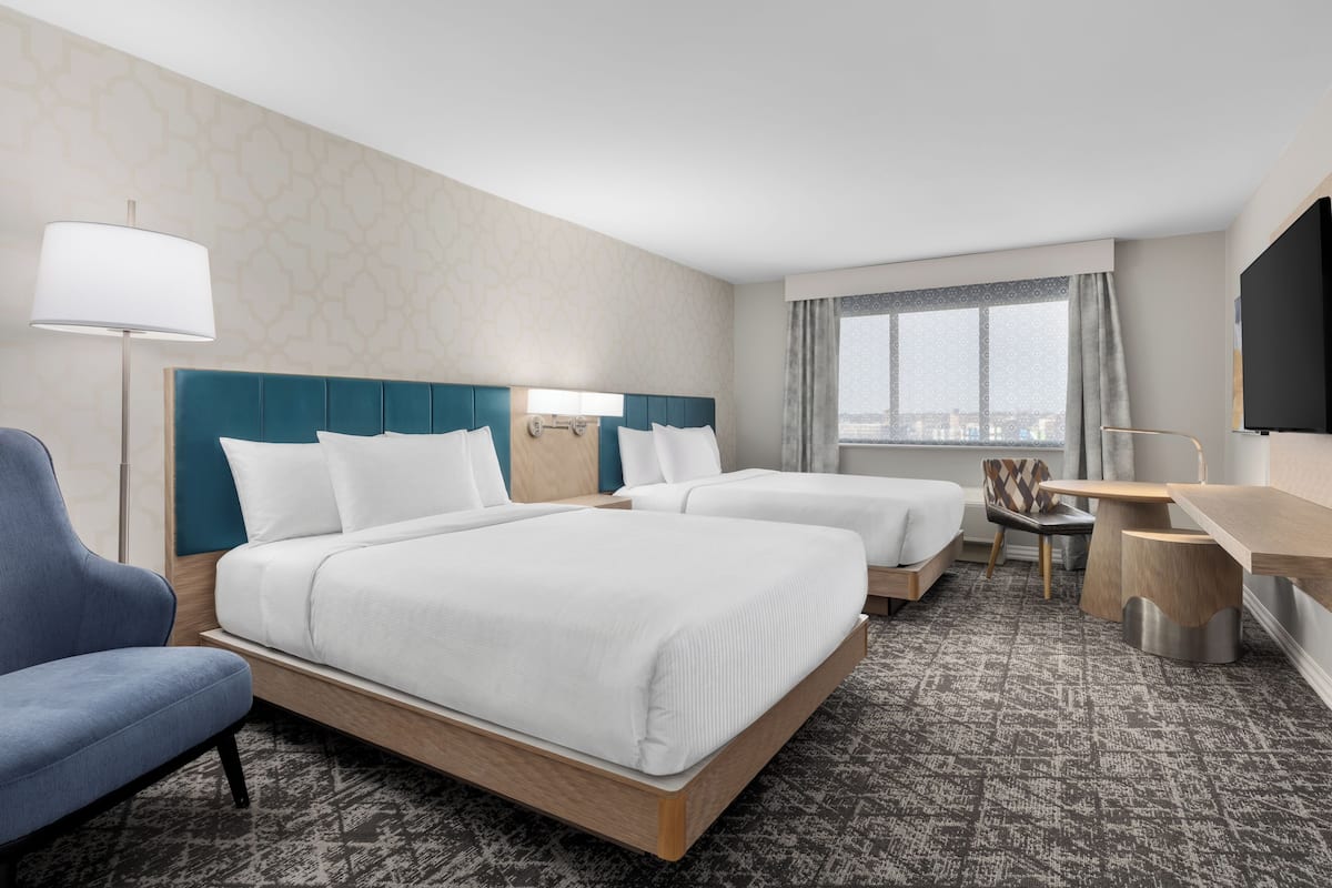 A hotel room with two beds, a desk, a chair, a TV, and a window with a city view.