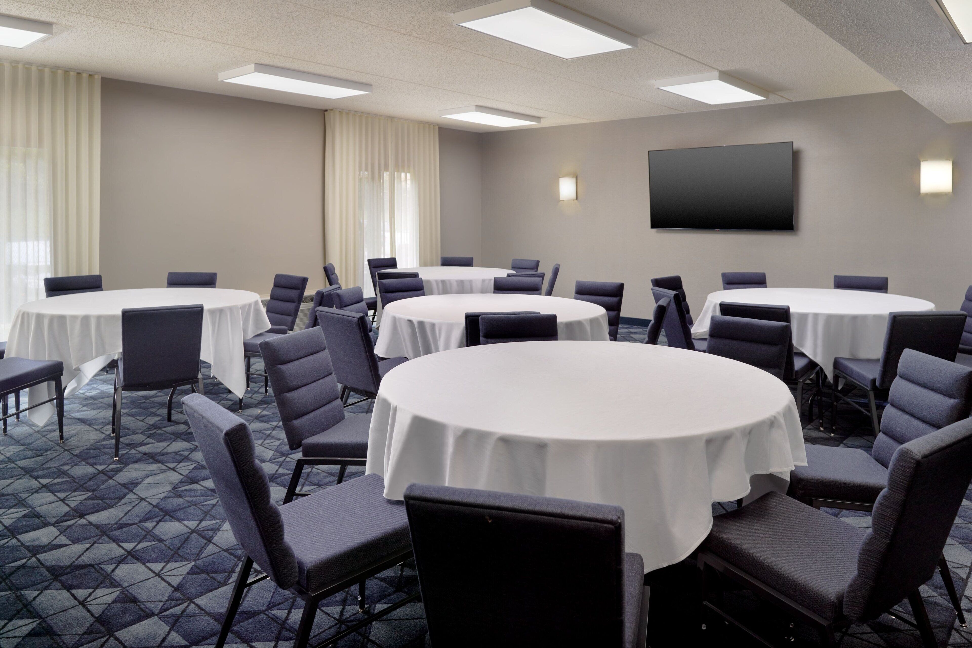 Une salle de conférence dotée de plusieurs tables rondes, chacune entourée de chaises, et d’un grand téléviseur à écran plat fixé au mur.