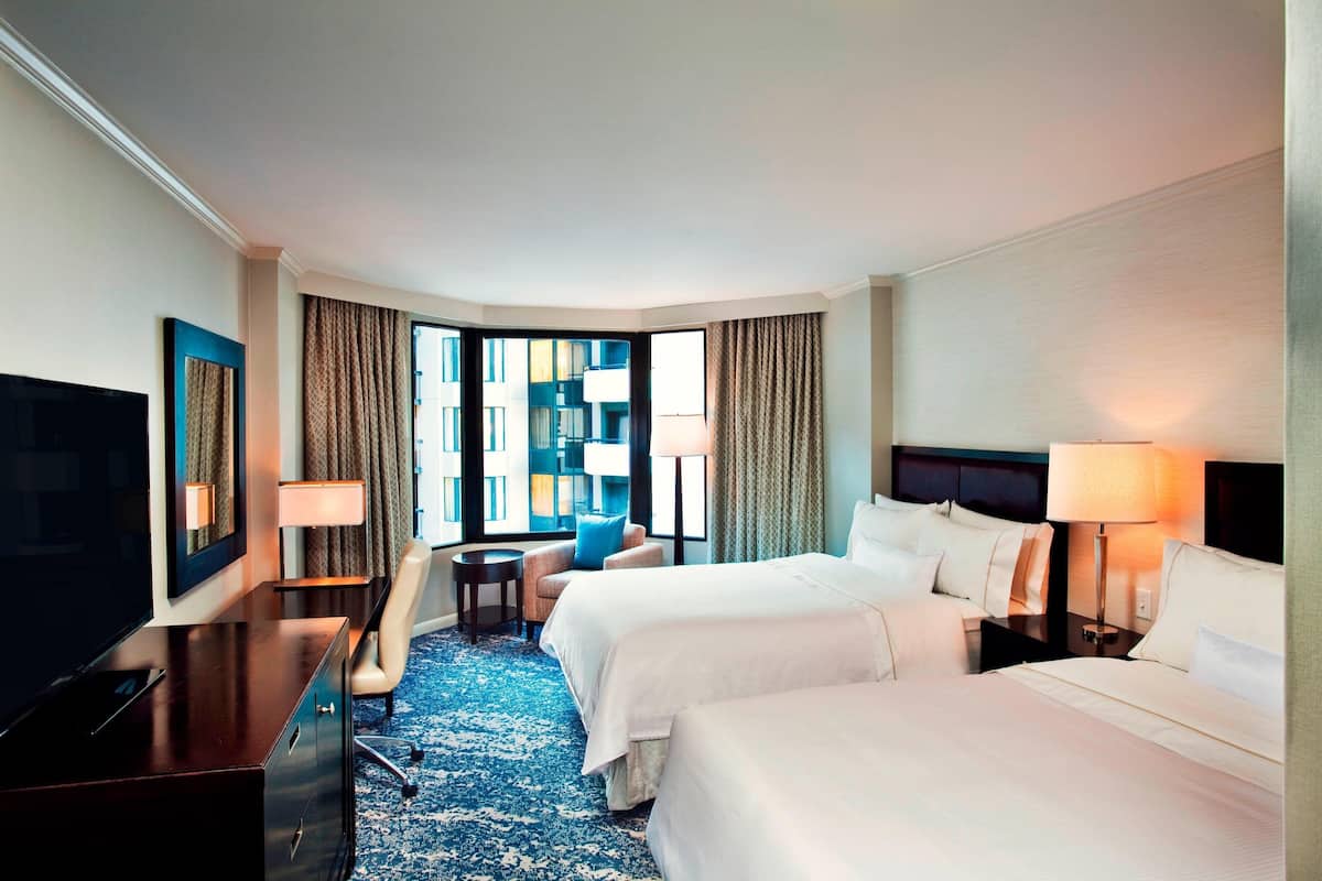 A hotel room with two beds, a desk, a TV, and a view of a building through large windows.