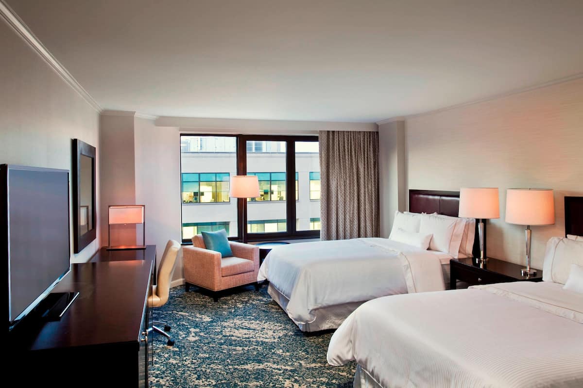A hotel room with two beds, a desk, a chair, a TV, and a large window with curtains.