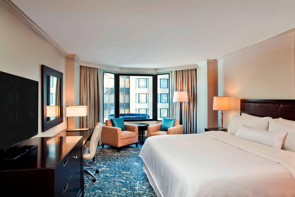 A hotel room with a large bed, a desk with a TV, two armchairs, a small table, and a view of buildings through large windows.