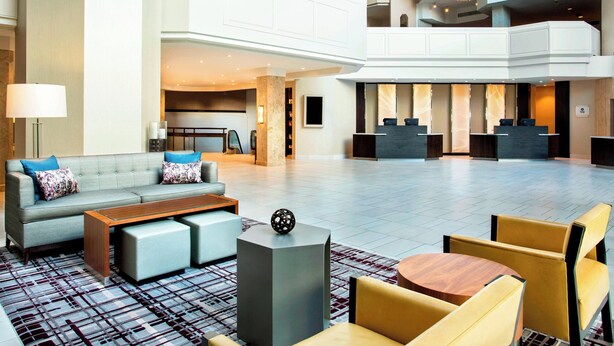 A spacious lobby with a grey sofa, a wooden coffee table, and yellow chairs. There is a reception desk with a black counter and a modern lamp.
