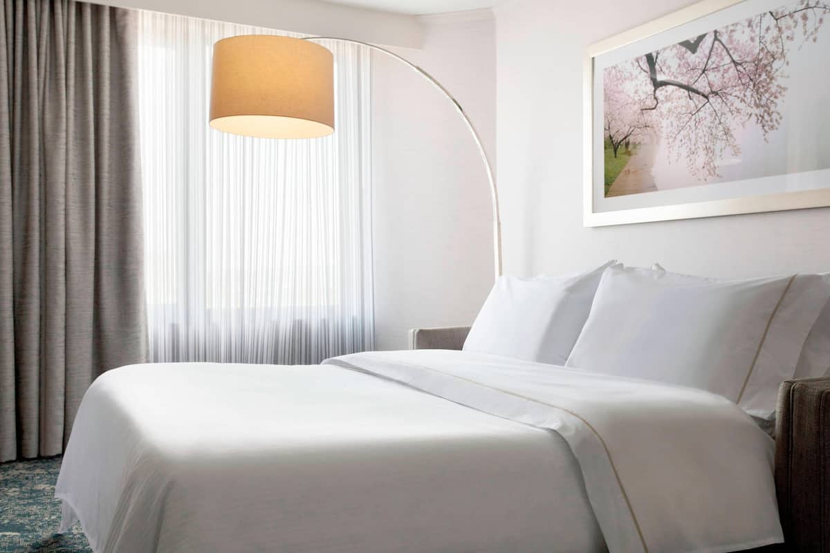 A hotel room with a large bed, a floor lamp, a window with sheer curtains, and a framed picture on the wall.