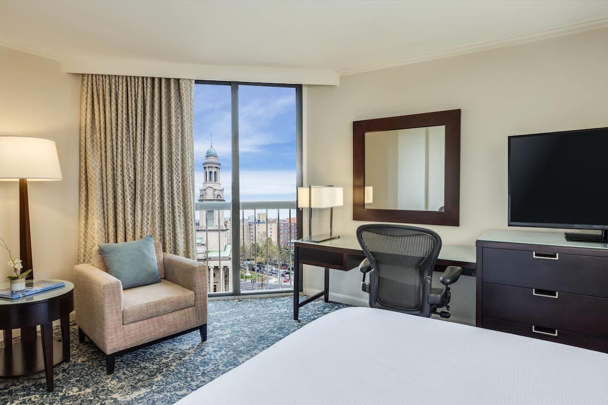 A hotel room with a large bed, a desk, a chair, a TV, a lamp, and a view of a building with a dome through the window.