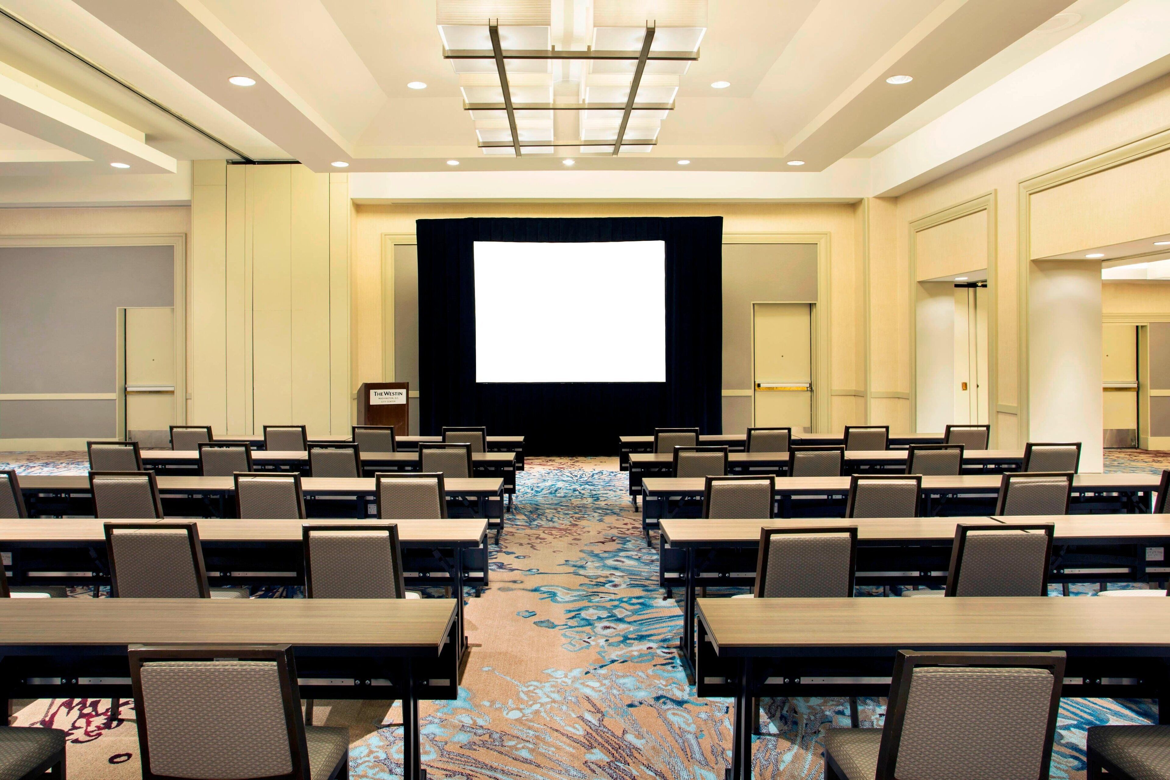 A conference room with rows of tables and chairs, a large screen at the front, and a carpeted floor.