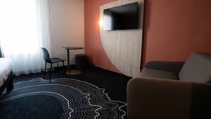 A hotel room with a bed, a sofa, a TV mounted on the wall, a small round table, and a chair. - ibis Styles Paris Porte de Versailles - Mairie d'Issy (Issy-les-Moulineaux)