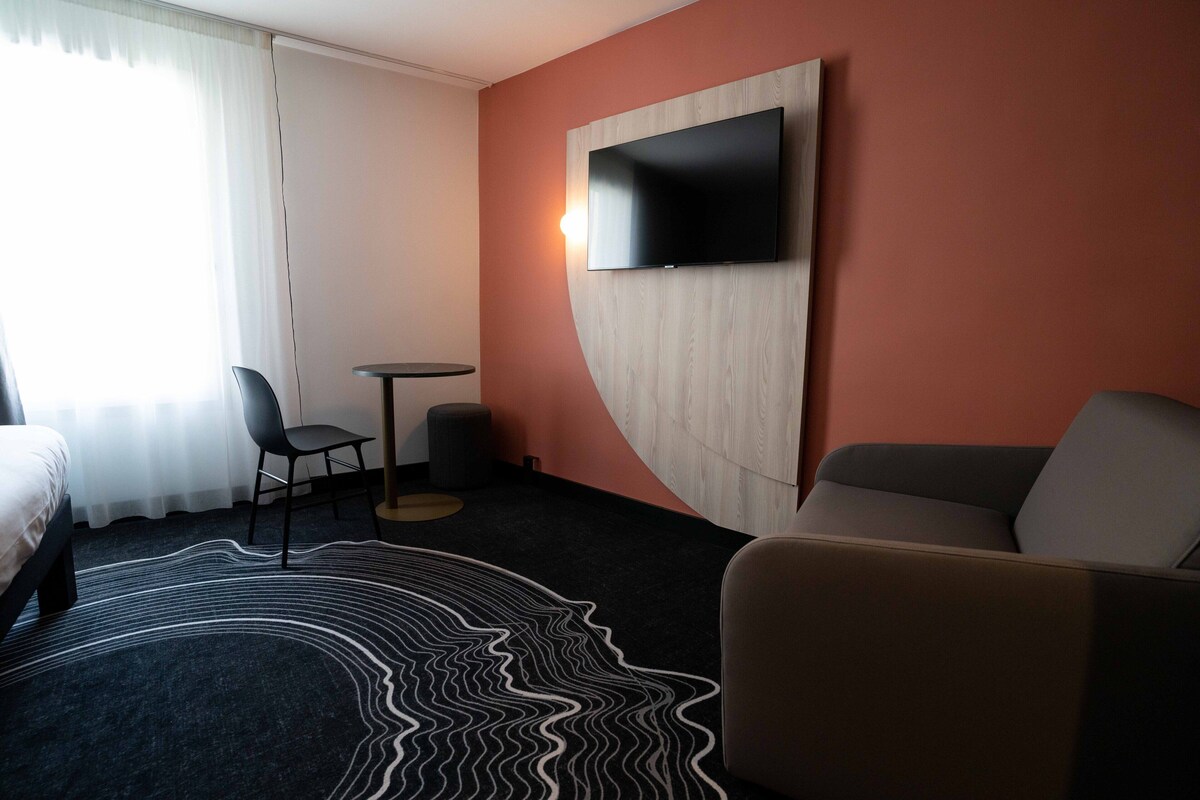 A hotel room with a bed, a sofa, a TV mounted on the wall, a small round table, and a chair.