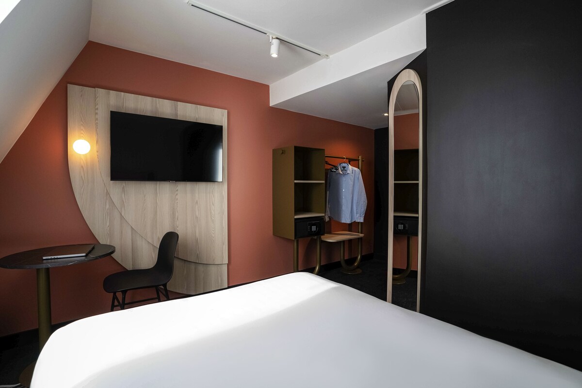 A modern hotel room with a large bed, a TV mounted on the wall, a small table, and a chair. There is a wardrobe with a mirror and a hanging rack.